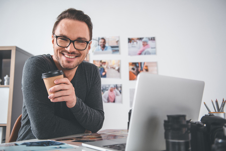 Portrait of cheerful bearded young man working with photos and digital device while sitting at table in apartment. He tasting mug of tea. Break conceptの写真素材