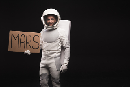 Hitchhike to planet. Portrait of spaceman wearing helmet with full armor is standing and looking at camera with smile. He is holding mars sign. Isolated background with copy space in the right sideの写真素材