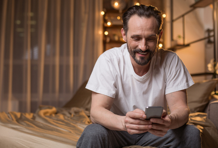 Portrait of happy male person messaging on cellphone at home. He is sitting on bed with relaxation and smiling. Copy spaceの写真素材
