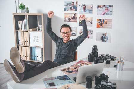 Full length portrait of cheerful unshaven photographer relaxing on chair near desk with camera, laptop and pictures. He flourishing arms. Relax conceptの写真素材