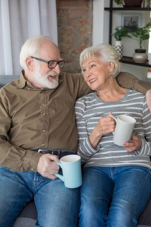 Happy time together. Portrait of aged smiling man and woman sitting on couch at home enjoying tasty teaの写真素材