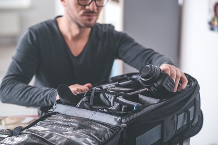 Close up male hand putting camera into big bag with other gadgets for images. Profession conceptの写真素材