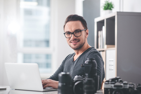 Portrait of cheerful young photographer typing in laptop while sitting at table. He looking at camera. Job conceptの写真素材