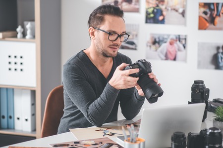Portrait of beaming unshaven young photographer holding digital device for creating pictures in arm while looking at it. Occupation conceptの写真素材