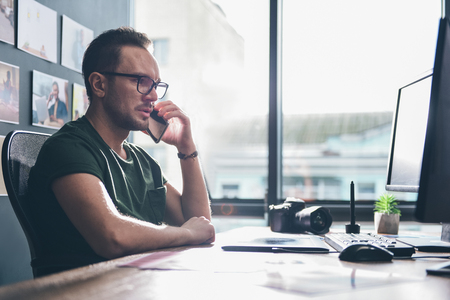 Side view orderly bearded photographer speaking by mobile while sitting at table and watching at screen of computer. Communication conceptの写真素材