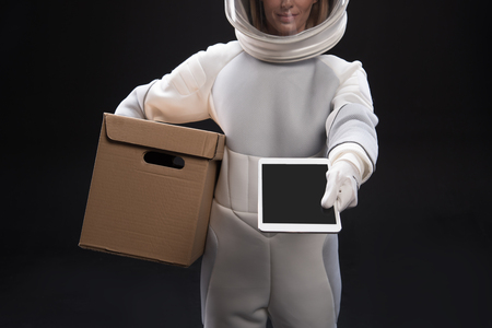 Sign it here. Pleasant spacewoman courier wearing helmet and protective suit is standing and keeping cardboard box while holding out digital tablet in hand. Selective focus. Space delivery conceptの写真素材