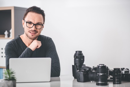 Portrait of smiling bearded man having work at digital device while locating at table. Labor concept. Camera equipment on table. Copy spaceの写真素材