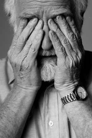 Close up portrait of sad pensioner expressing depression while standing. He is closing face with palmsの写真素材