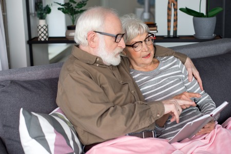 Side view of two old man and woman. They are remembering sweet moments while resting on comfortable couch at home. Both are pointing with finger at book while husband is cuddling wifeの写真素材