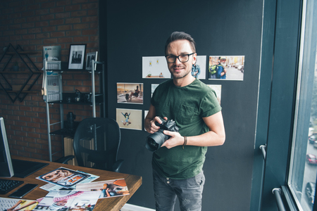 Portrait of smiling unshaven photographer holding camera in arms while locating in modern apartment. Occupation and hobby conceptの写真素材