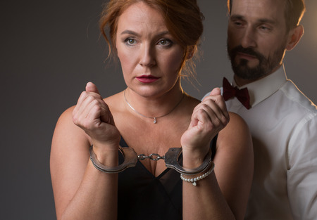 I am your prisoner. Portrait of frustrated middle-aged woman showing her hands locked by handcuffs. Man is standing near her and looking at lady with confidence. Isolatedの写真素材