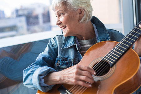Waist up portrait of charming female pensioner playing guitar at home and smilingの写真素材
