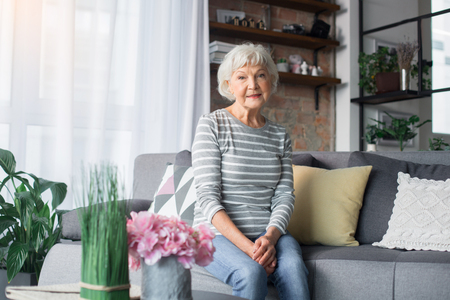 Portrait of mature woman resting on comfortable couch at home. She is smiling and looking at cameraの写真素材