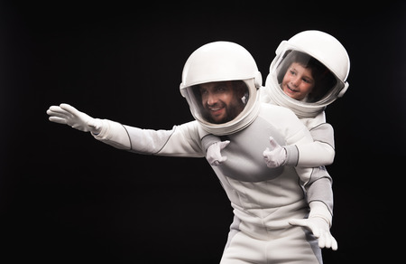 Always fun. Cheerful father and little son wearing hyperbaric astronaut protective suit and helmet are playing together. Boy is pretending to fly while sitting on back of man. Isolated backgroundの写真素材