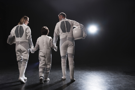Friendly relations. Back view of positive family of astronauts are going together while holding by hands. Mother and father are looking at their little son. Dark background with copy spaceの写真素材