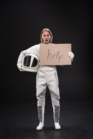 Full length portrait of frightened spacewoman wearing protective costume is standing and holding helmet and cardboard sign help. She is feeling consternation. Danger concept. Isolated backgroundの写真素材