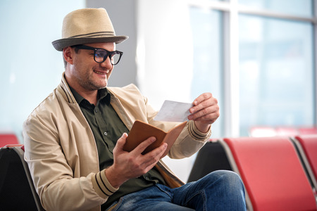 Checking details. Joyful middle-aged man in hat is looking at ticket with smile while sitting on bench at airport lounge. He is waiting for his flight. Copy space in the right sideの写真素材