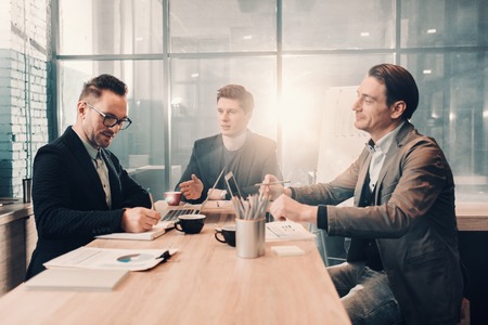 Portrait of smiling bearded man writing in copybook while speaking with happy partners. They situating at desk. Communication and business conceptの写真素材