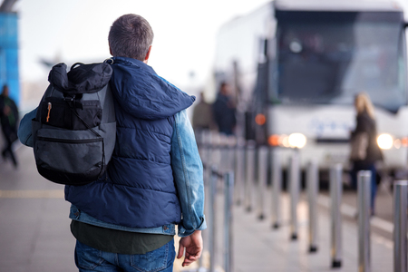 Modern tourist. Back view of mature man with backpack is standing near road of international airport. Bus stop in background. Copy space in the right sideの写真素材