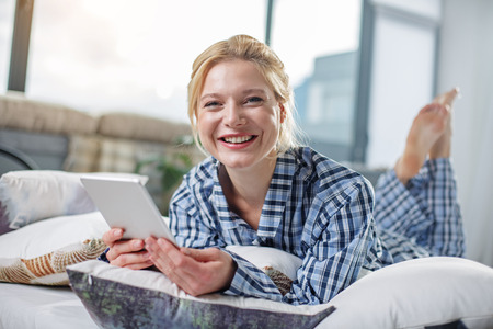 Portrait of laughing lady lying on home bed with gadget in hands. She is looking at camera with...