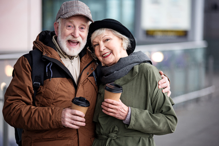 Together forever. Waist up portrait of happy aged couple is standing together outdoors and holding coffee. Men is hugging elegant woman and expressing admiration. They are looking at camera with joyの写真素材