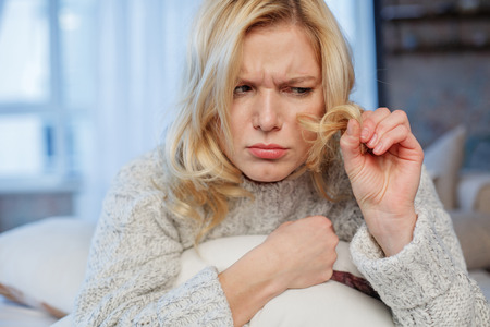 Portrait of puzzled girl sitting at home. She is holding lock of hair and looking at it with sadnessの写真素材