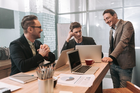Portrait of cheerful man looking at screen of laptop of partner while standing near him. Cheerful bearded affiliate watching at them. Profession conceptの写真素材