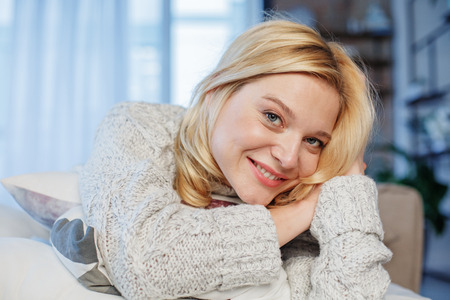 Portrait of beautiful lady looking at camera with coquetry and showing wide smile. She is sitting in living roomの写真素材