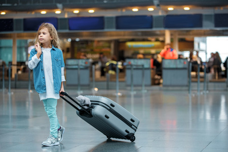 First journey. Full length of stylish cute blond child is standing at airport building and carrying her suitcase with toy bear. She is looking aside pensively while waiting for flight. Copy spaceの写真素材