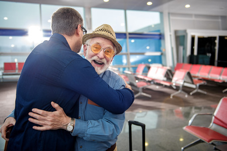 Happy moment. Portrait of pleased aged father in glasses and his grownup child are hugging each other while standing at airport lounge. Old man is looking at camera with glad. Copy space in right sideの写真素材