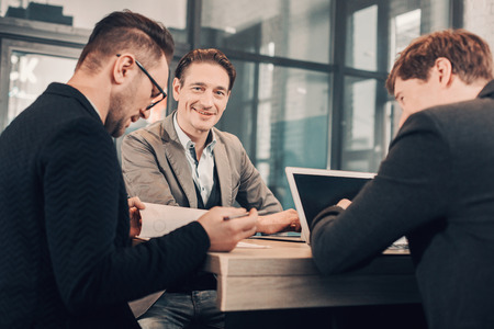 Portrait of cheerful young male telling with happy colleagues while sitting at table in apartment. Conversation and job conceptの写真素材