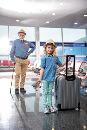 My first travel. Full length portrait of cute little girl is holding suitcase while looking at camera with joy. Her pleasant aged bearded man is resting on walking stick in background. Selective focusの写真素材