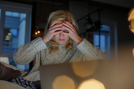 Portrait of blond girl on sofa with laptop. She is covering her eyes with hands and looking disturbedの写真素材