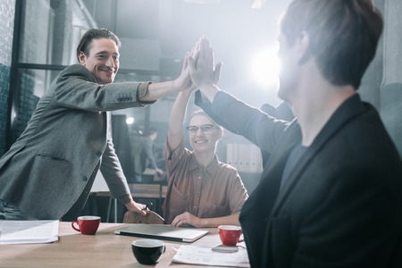 Low angle happy males and cheerful woman rising hands into pyramid while locating at desk in room....