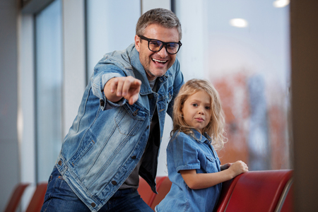 Feeling curious. Cheerful mature father and his small daughter are sitting on airport seats. Parent is pointing finger on camera with joy while girl is looking there with slight smileの写真素材