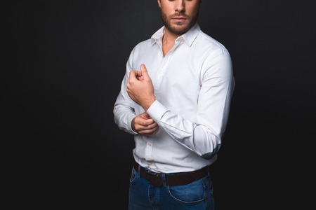 Untroubled well-dressed man standing and fixing the button on handcuff. Isolated on black background. Copy space in left sideの写真素材