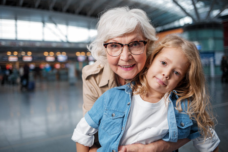 Happy moment. Portrait of charming positive gray-haired grandmother is hugging her pretty granddaughter from behind. They are looking at camera with joy while standing at airport hall. Copy spaceの写真素材