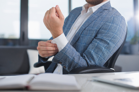 Always good looking. Fashionable clerk sitting on office chair and buttoning on cuff. Close up of his armの写真素材