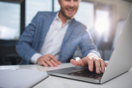 Smiling businessman sitting in the office and typing on a computer. Focus on deviceの写真素材