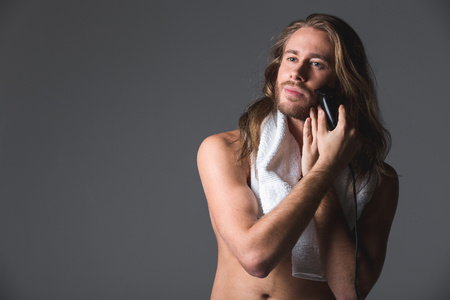 Waist up portrait of peaceful naked guy shaving his cheek with white towel on his neck. Isolated on grey background. Copy space in left sideの写真素材