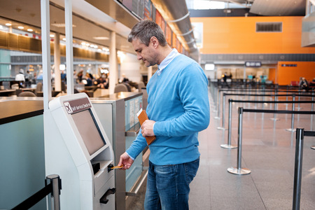 Side view profile of serious mature man is standing near self-service check-in kiosk at international airport building. He is using his credit card to making payment for flight ticketsの写真素材