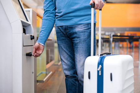 Close-up of male arm is pushing credit card to self-service check-in kiosk to making payment for flight tickets. He is standing at international airport building while holding suitcaseの写真素材