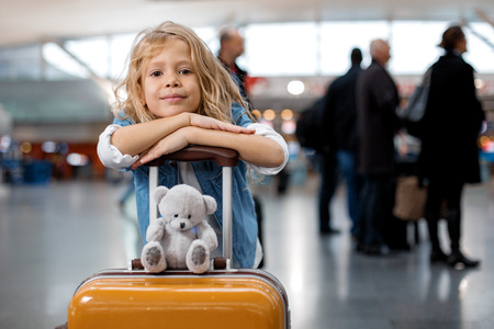 Travel with friend. Portrait of joyful stylish female child is leaning on handle of suitcase with toy bear at airport lounge. She is looking at camera with joy. Copy space with people in backgroundの写真素材