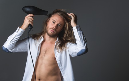 Waist up portrait of calm man holding hairdryer and looking at camera. His shirt is unbuttoned. Copy space in right side. Isolated on grey backgroundの写真素材