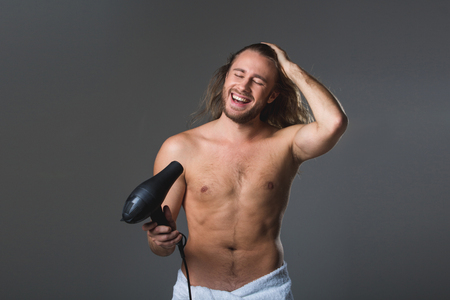 Waist up portrait of longhaired satisfied guy with bare torso holding hairdryer. His eyes are closed. Isolated on grey backgroundの写真素材