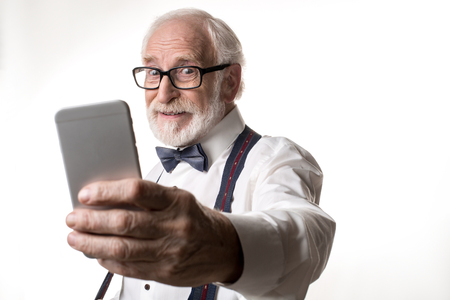 Waist up portrait of joyful elderly man making selfie, looking amused. Focus on face and phone. Isolated on backgroundの写真素材