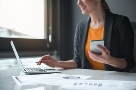 Close up female hand keeping phone while typing in modern laptop in office. Technology and job conceptの写真素材