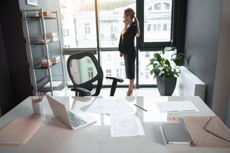 Full length side view serene businesswoman telling by mobile while situating near window in office. Conversation and job concept. Focus on table with documents and laptopの写真素材