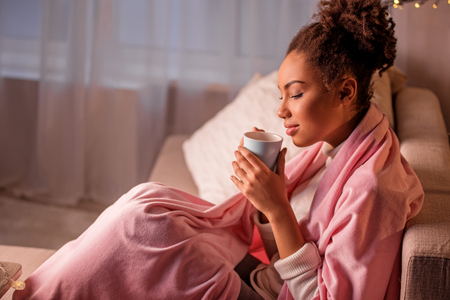 Serene african girl is drinking tea with pleasure. She is sitting on couch at home in cozy atmosphereの写真素材
