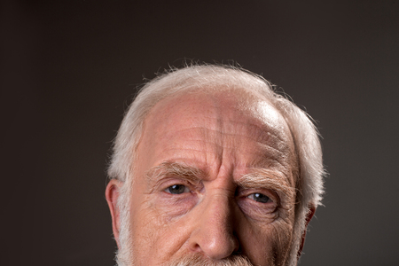 Close up of gray haired male half face wrinkling his forehead, expressing tiredness. Isolated on grey backgroundの写真素材